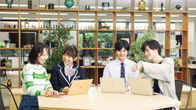 4人の学生が机を囲み、それぞれノートパソコンを使いながら楽しそうに話し合っている様子