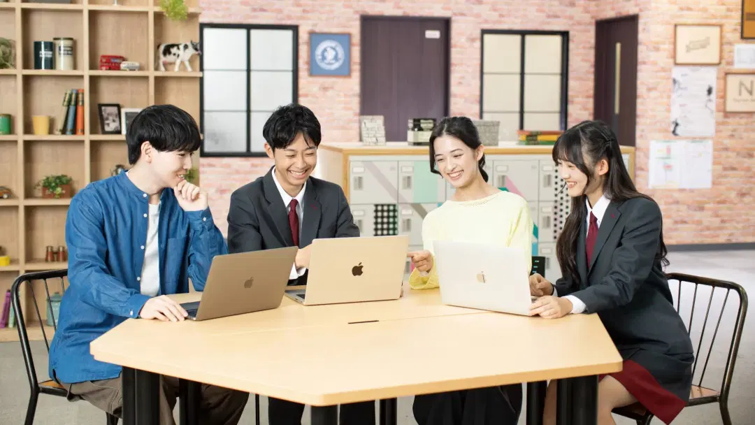4人の学生が机を囲み、それぞれノートパソコンを使いながら楽しそうに話し合っている様子